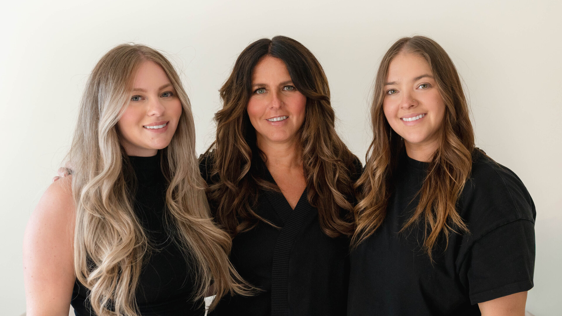 The founders of Momé — a mother and her two daughters — standing together in black, smiling, against a soft neutral wall.