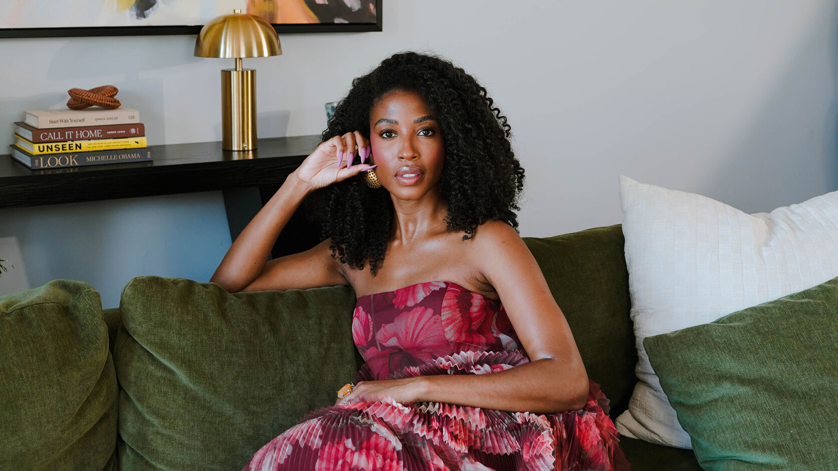 Aisha Beau Frisbey, in a strapless pleated magenta floral gown, sits on a green velvet sofa with one hand raised to her ear, beneath an abstract painting beside a brass lamp and a stack of books.