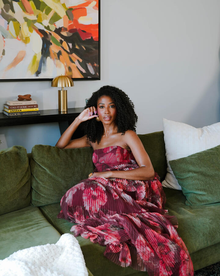 Aisha Beau Frisbey, in a strapless pleated magenta floral gown, sits on a green velvet sofa with one hand raised to her ear, beneath an abstract painting beside a brass lamp and a stack of books.