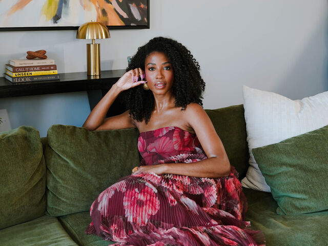 Aisha Beau Frisbey, in a strapless pleated magenta floral gown, sits on a green velvet sofa with one hand raised to her ear, beneath an abstract painting beside a brass lamp and a stack of books.