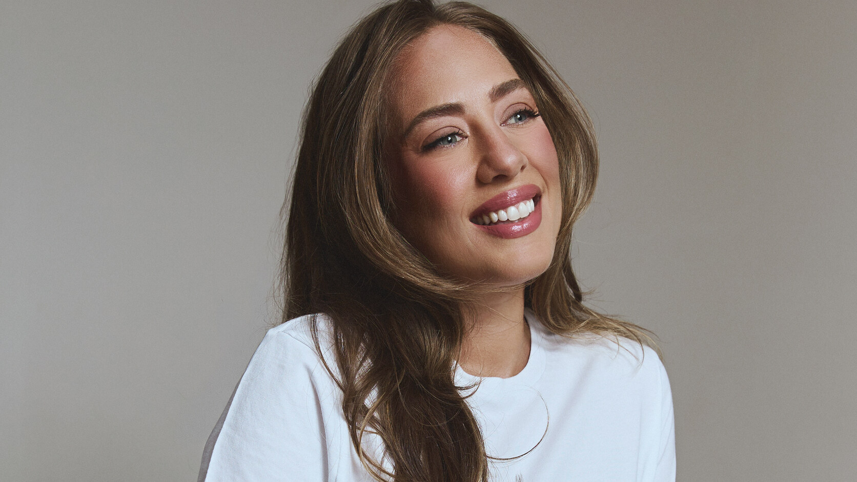 Amanda Hirsch sits on a stool in a white t-shirt and dark jeans against a warm grey backdrop, smiling and looking off camera.