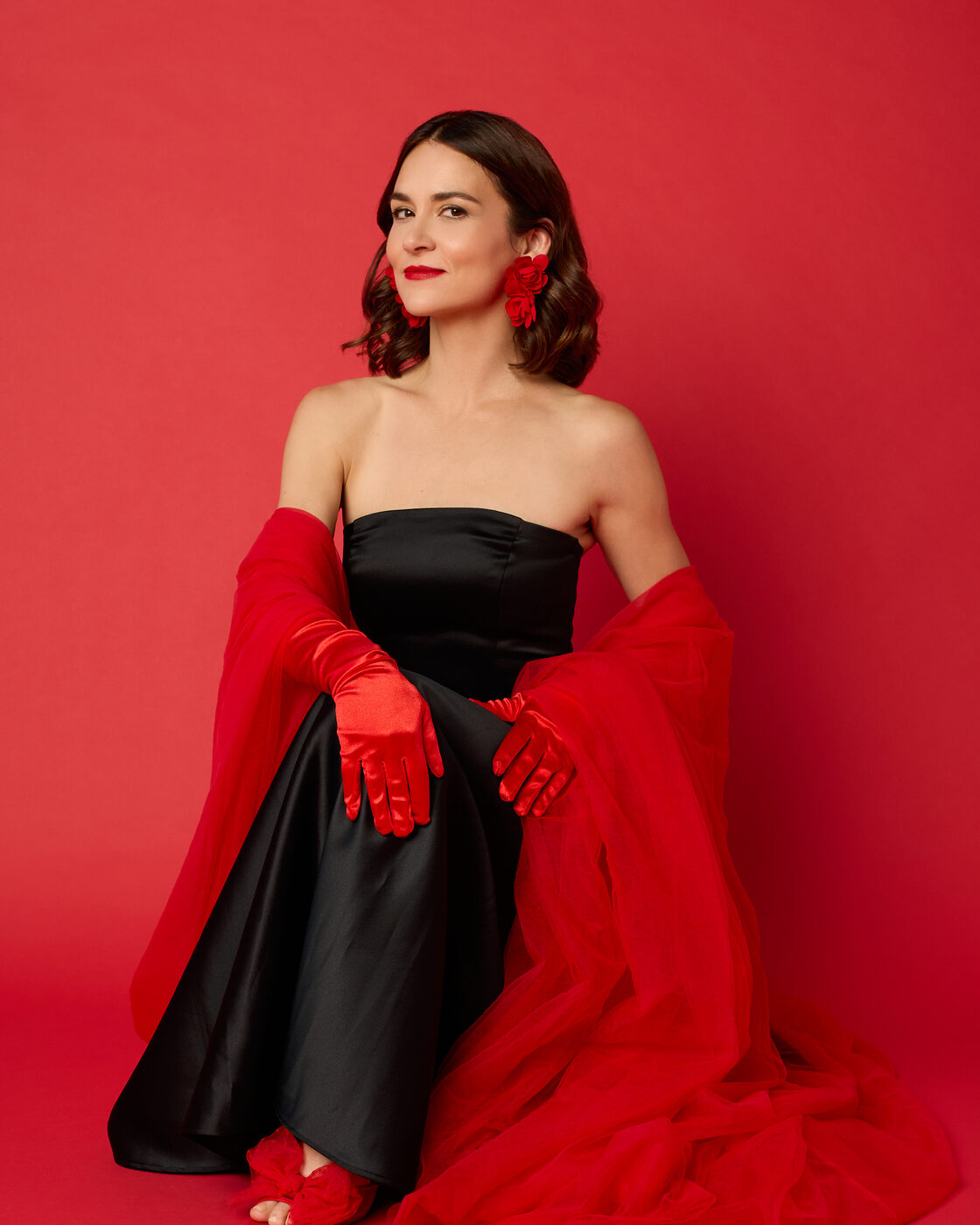 Ava Paloma sits against a red backdrop in a black strapless gown with long red satin gloves, red floral earrings and a red tulle wrap.