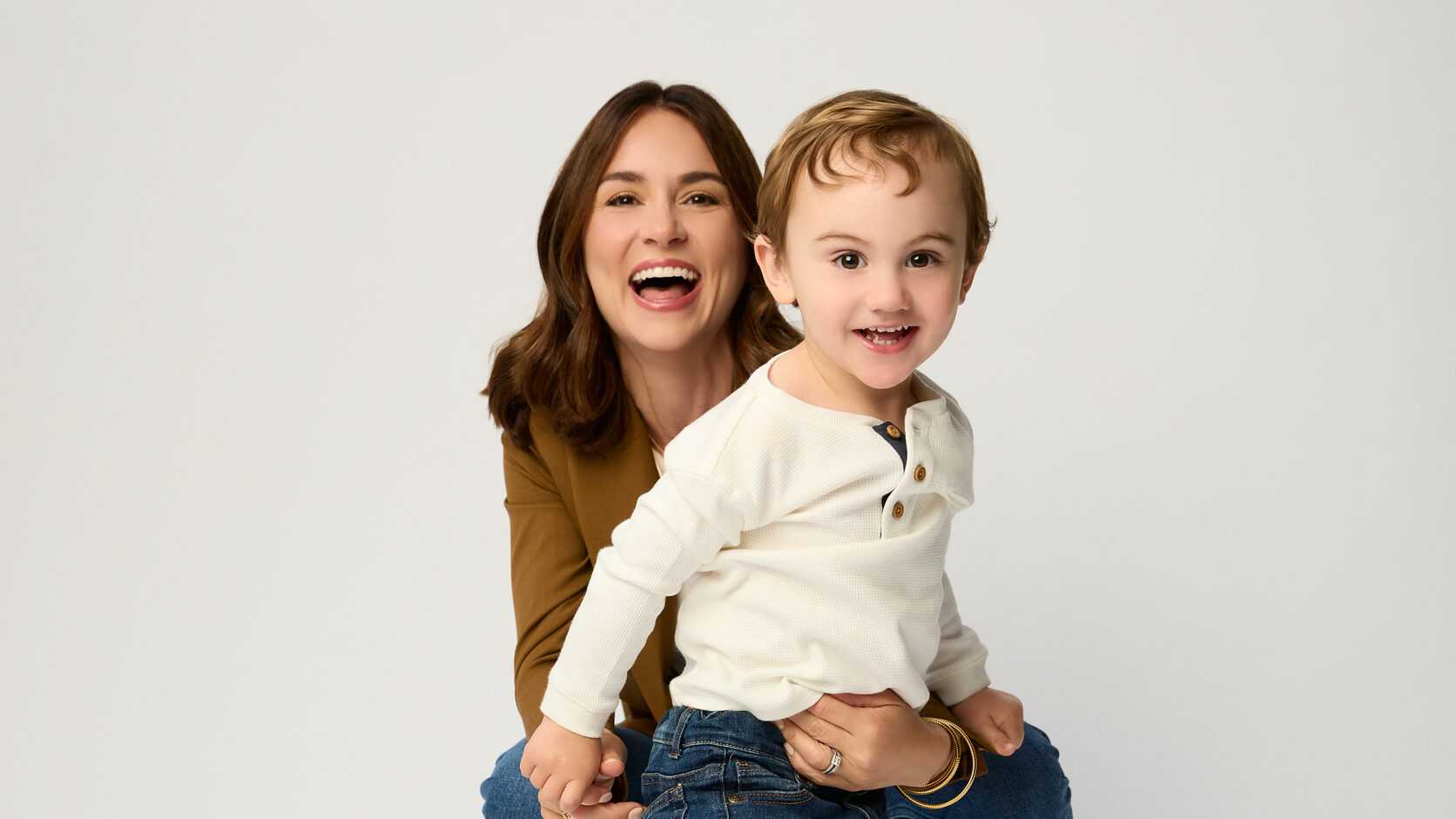 Ava Paloma laughs as she crouches behind her toddler son Bo, who stands in front of her in a cream henley and jeans, against a white studio backdrop.