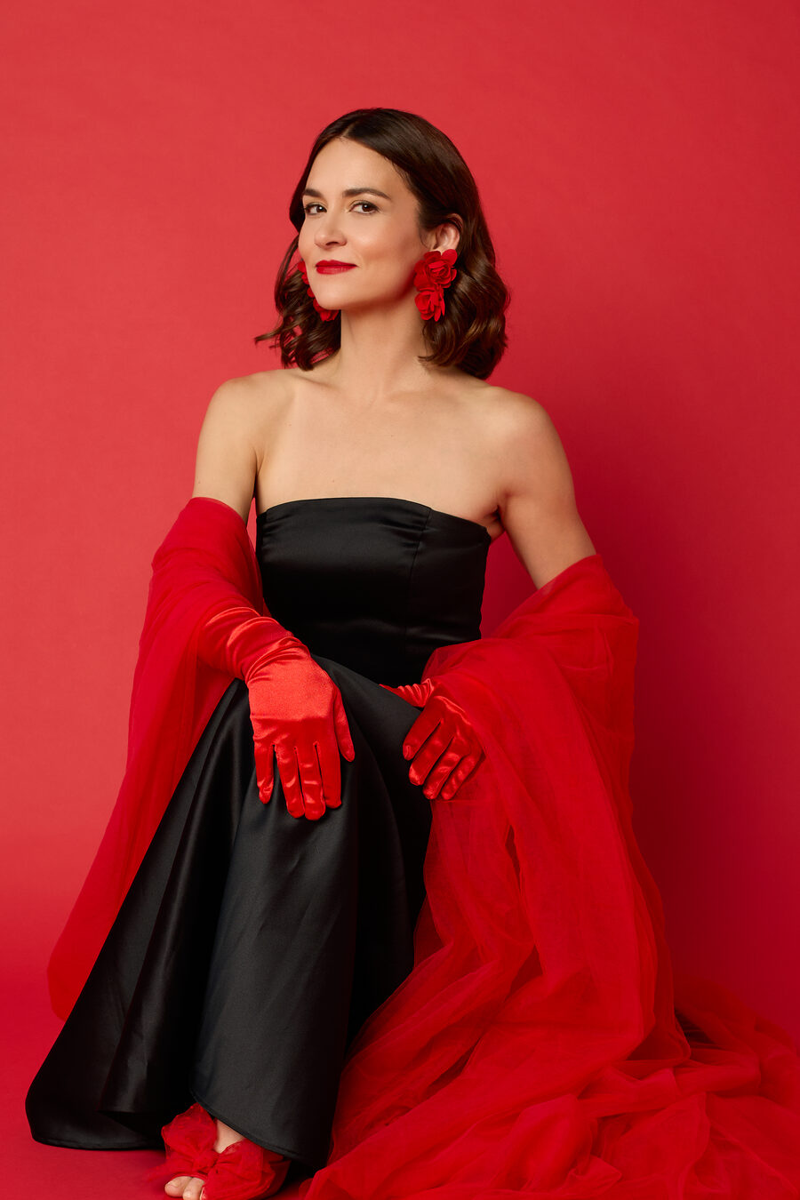 Ava Paloma sits against a red backdrop in a black strapless gown with long red satin gloves, red floral earrings and a red tulle wrap.