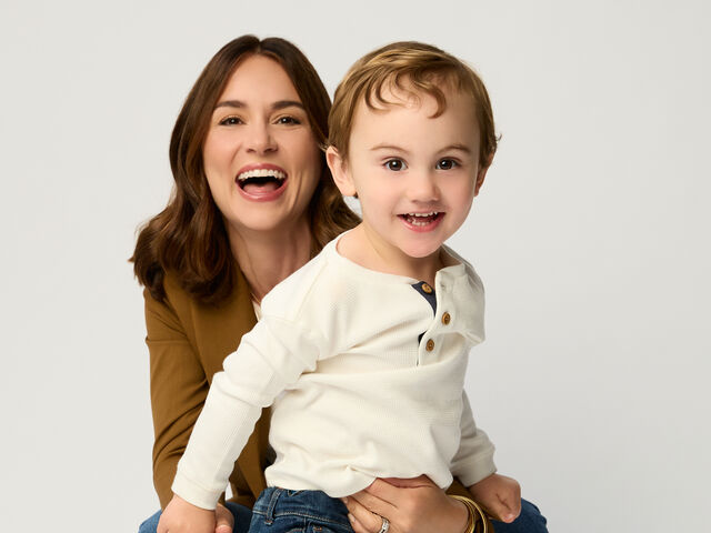 Ava Paloma laughs as she crouches behind her toddler son Bo, who stands in front of her in a cream henley and jeans, against a white studio backdrop.