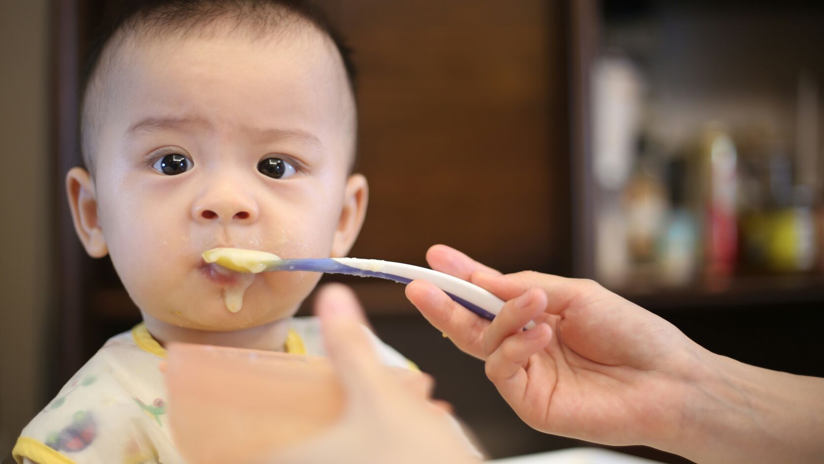 A baby in a high chair is spoon-fed a smooth yellow puree, a little of it on their chin.