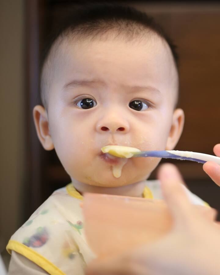 A baby in a high chair is spoon-fed a smooth yellow puree, a little of it on their chin.