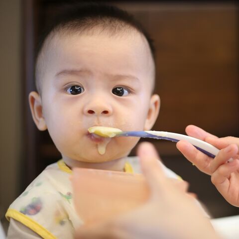 A baby in a high chair is spoon-fed a smooth yellow puree, a little of it on their chin.