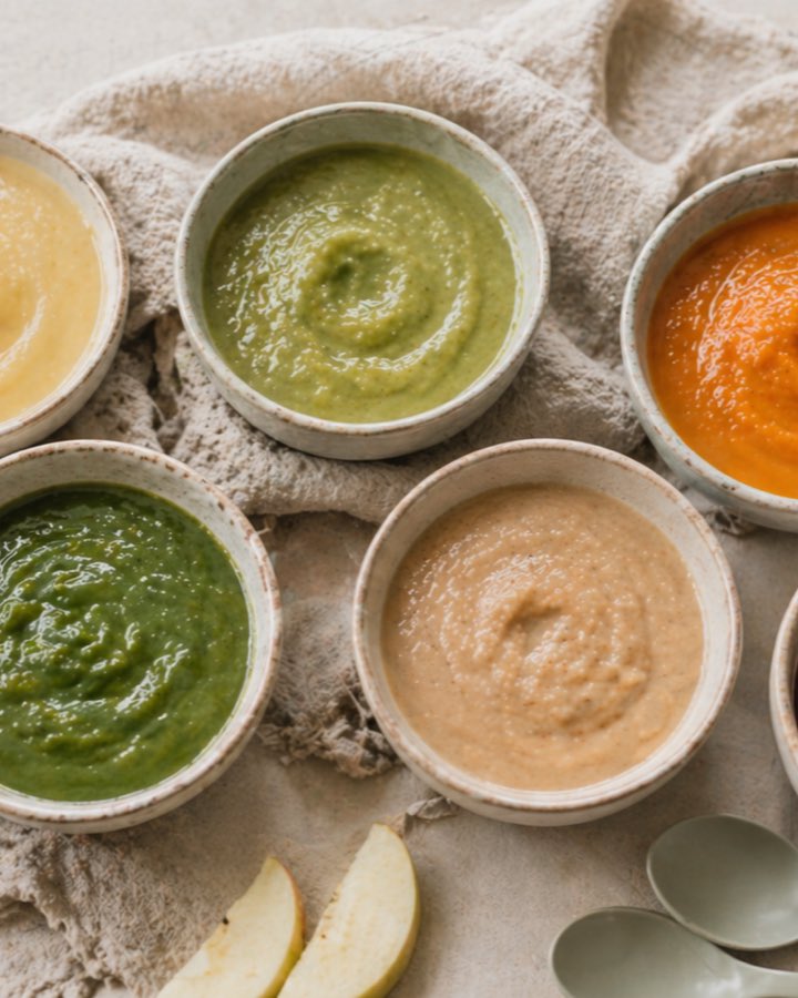 Six small ceramic bowls of smooth, brightly coloured homemade baby purees on a linen cloth, surrounded by fresh vegetables and fruit.