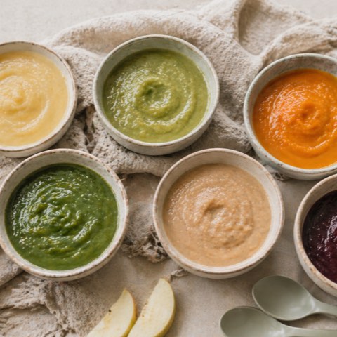 Six small ceramic bowls of smooth, brightly coloured homemade baby purees on a linen cloth, surrounded by fresh vegetables and fruit