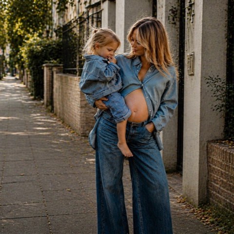 A mother in wide-leg denim holds her toddler on a sunlit, tree-lined street, both of them in soft golden afternoon light.