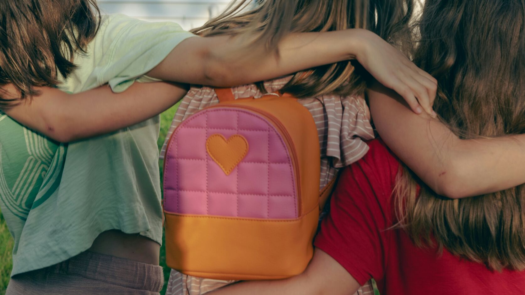 Three children stand with their arms around each other, seen from behind, one wearing a small quilted pink and orange backpack with a heart patch.