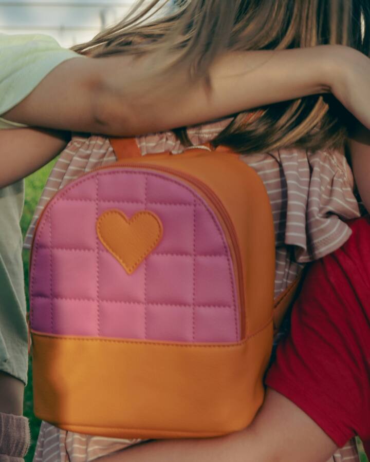 Three children stand with their arms around each other, seen from behind, one wearing a small quilted pink and orange backpack with a heart patch.