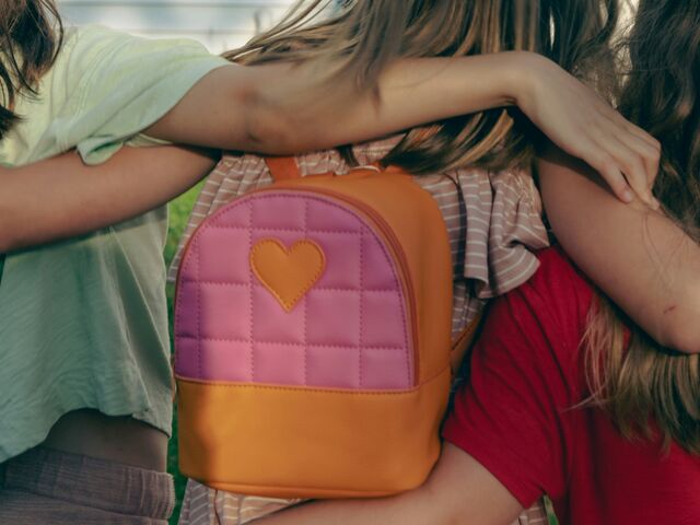 Three children stand with their arms around each other, seen from behind, one wearing a small quilted pink and orange backpack with a heart patch.