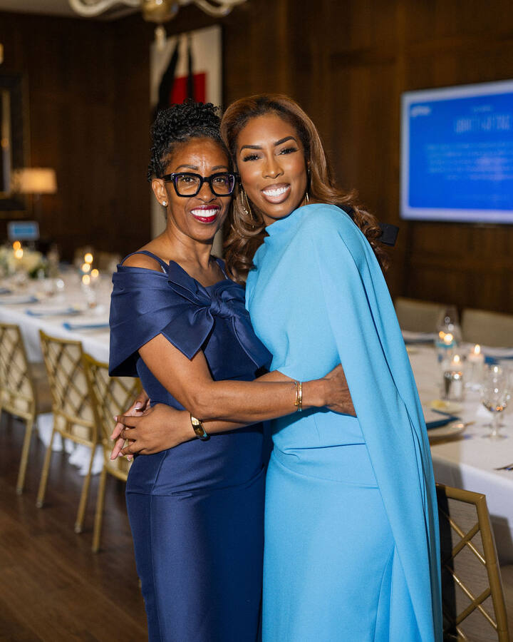 Beverly Beal Sr., in black-framed glasses and an off-the-shoulder navy satin dress, hugs her daughter Beverly Beal Jr., in a sky-blue caped gown, as they smile at the camera in a wood-panelled dining room set for dinner.