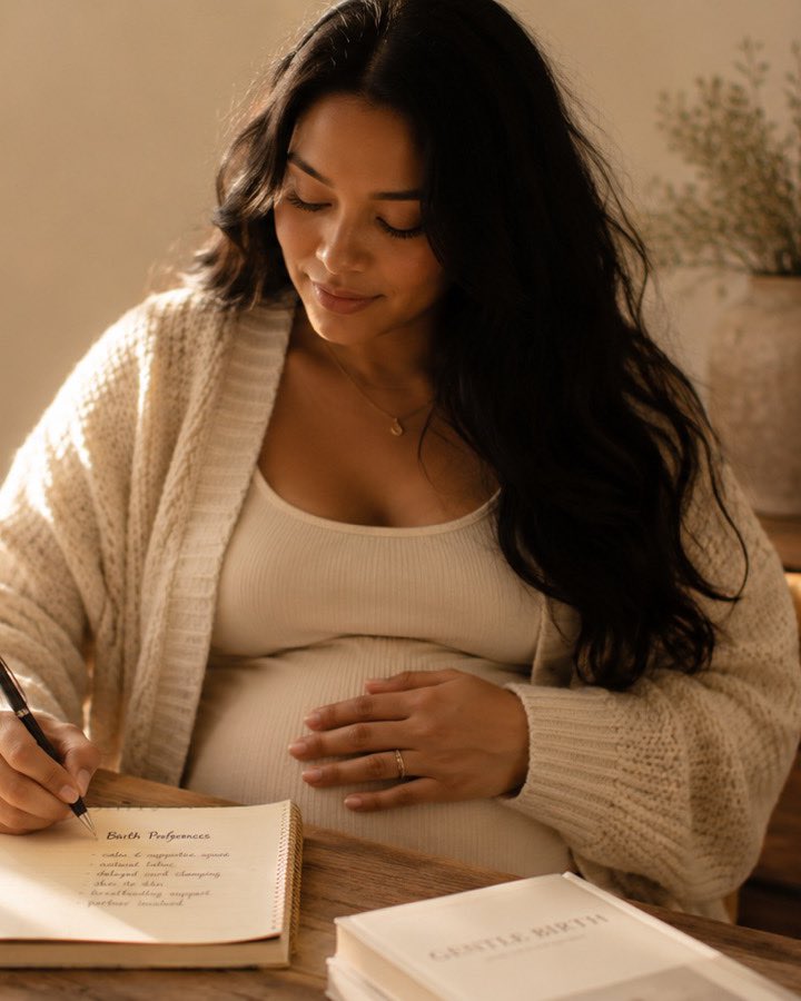 A pregnant woman in a cream slip dress sits at the window of a soft, sunlit room, one hand resting on her belly.