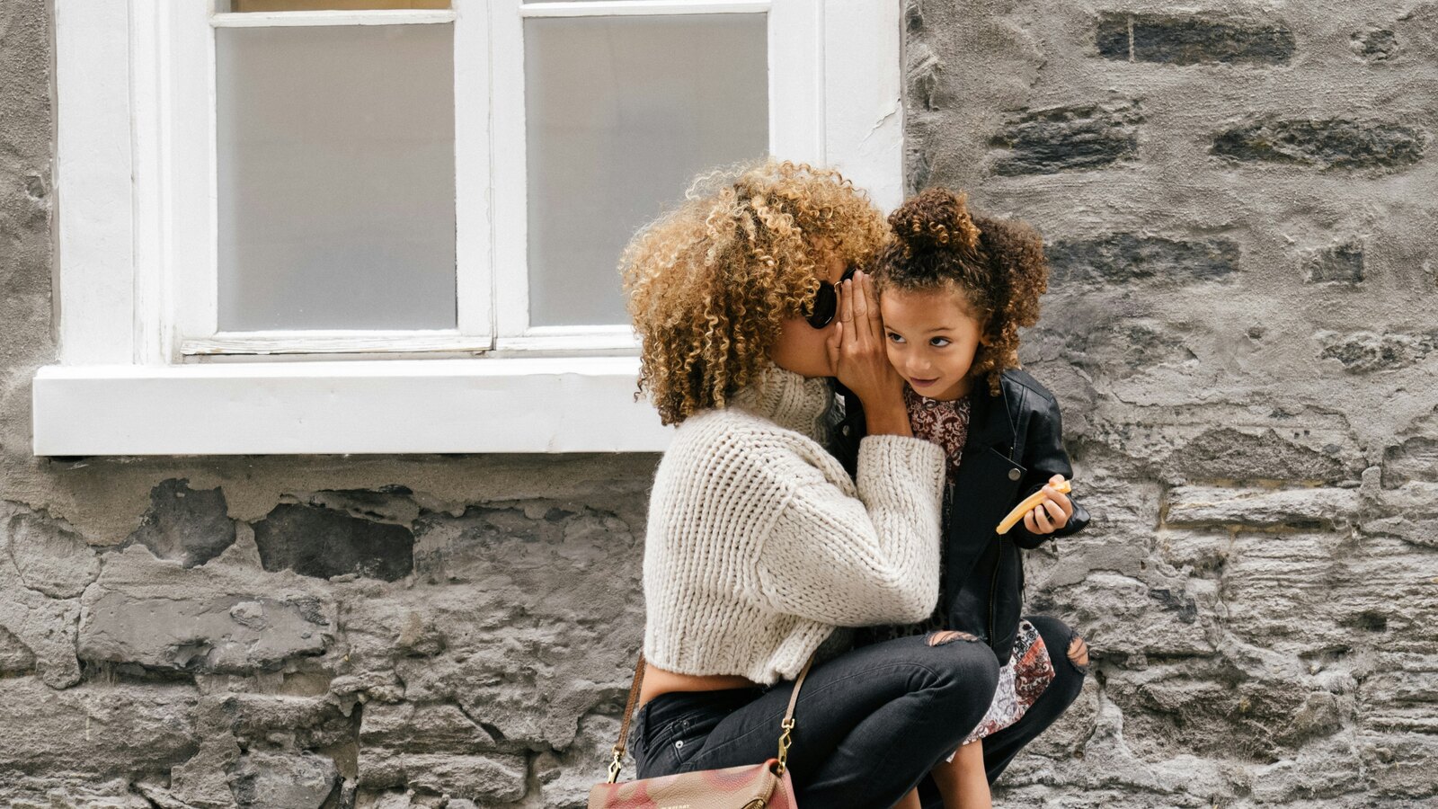 A mother with curly hair leans in to whisper to her young daughter as they crouch together against a stone wall on a city street.