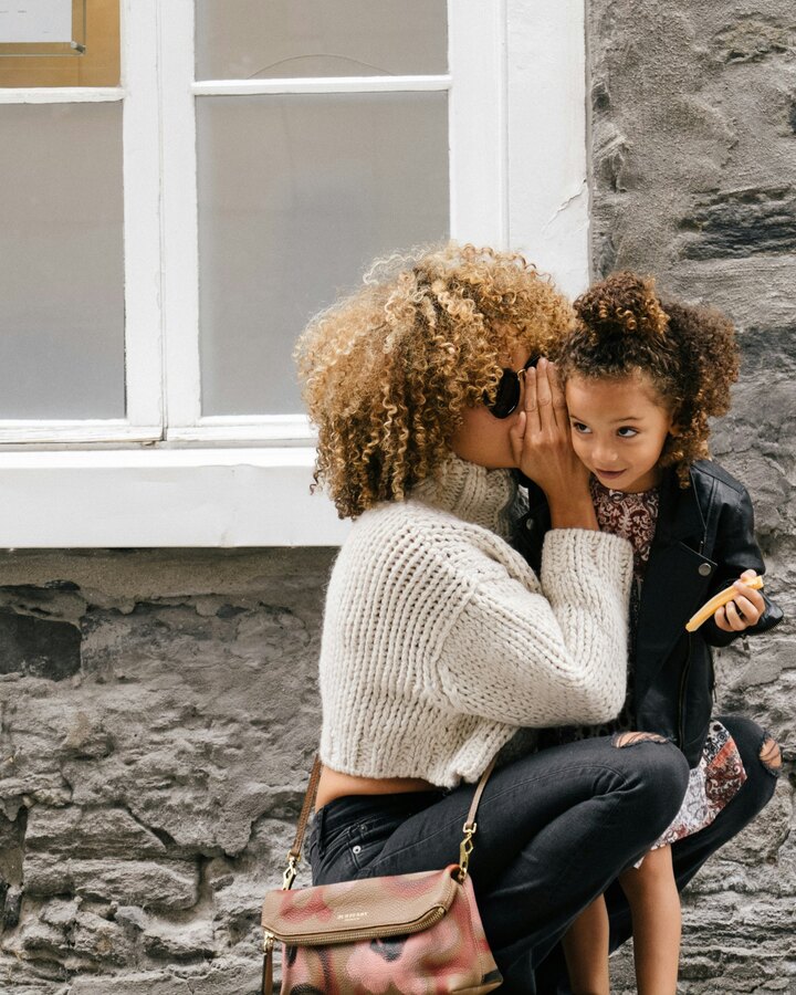 A mother with curly hair leans in to whisper to her young daughter as they crouch together against a stone wall on a city street.