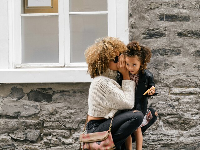 A mother with curly hair leans in to whisper to her young daughter as they crouch together against a stone wall on a city street.