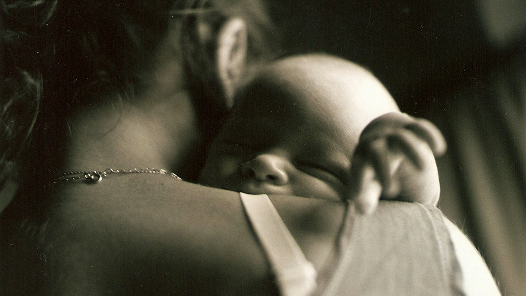 A mother holds her sleeping newborn against her shoulder in soft sepia light, the baby's small hand resting on her back.