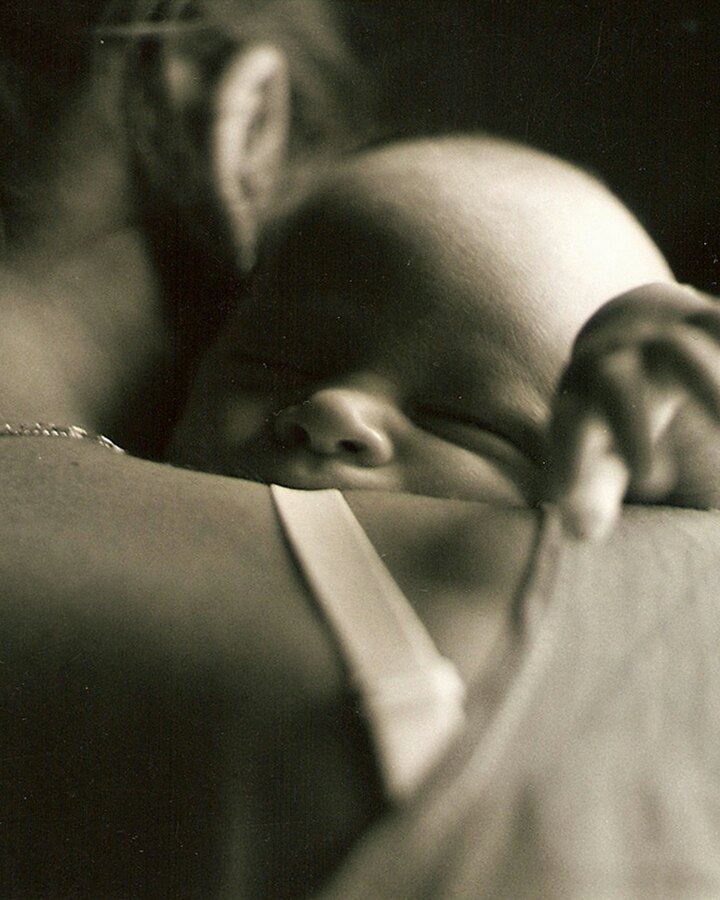 A mother holds her sleeping newborn against her shoulder in soft sepia light, the baby's small hand resting on her back.