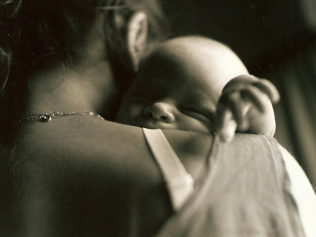 A mother holds her sleeping newborn against her shoulder in soft sepia light.
