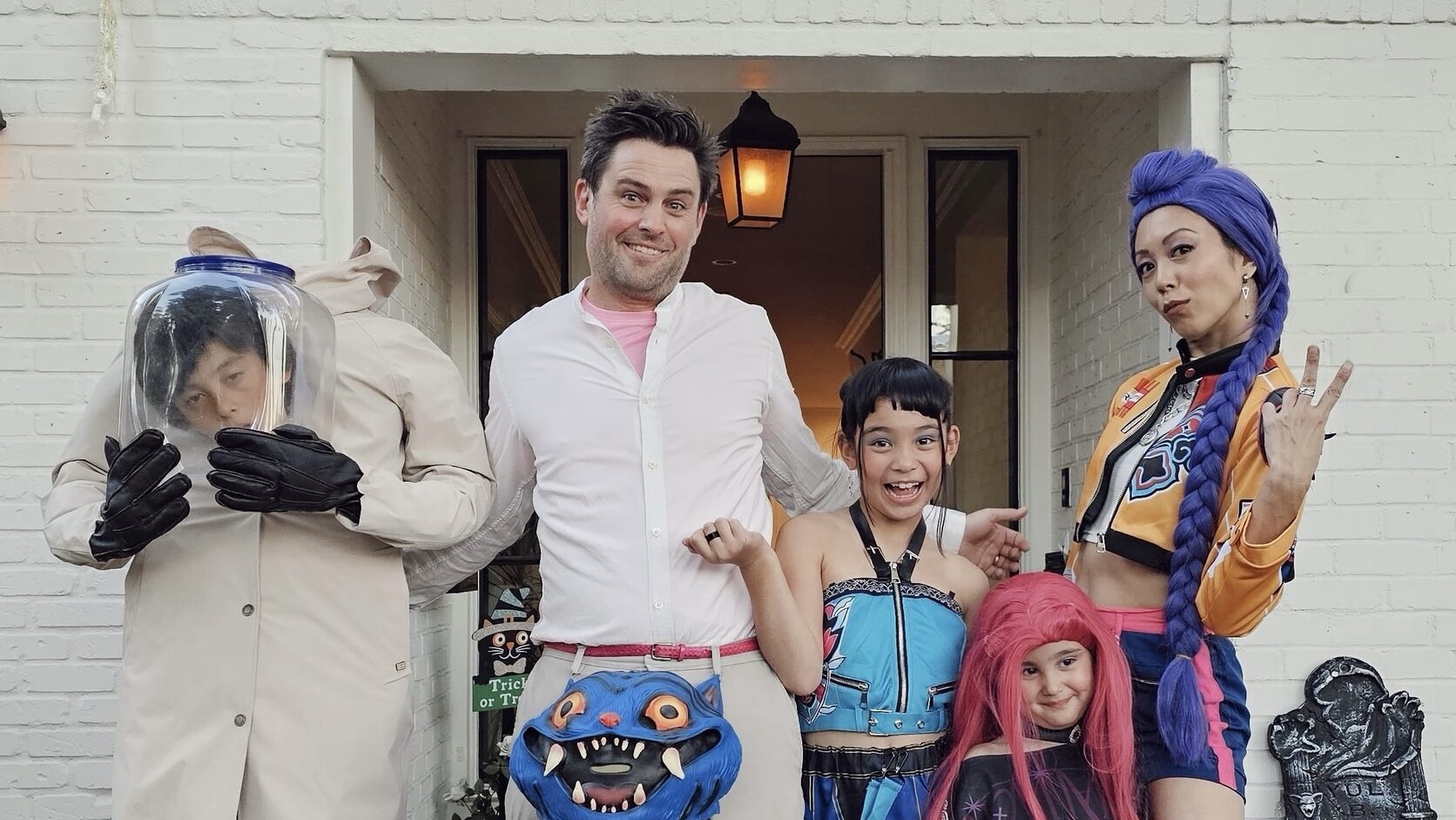 Brittany Ishibashi, her husband, and their three children pose in Halloween costumes outside their white brick front door — she in a long blue braided wig and orange jacket flashing a peace sign, he in a blue monster costume, the kids in a fishbowl helmet, a blue anime outfit, and a pink wig.