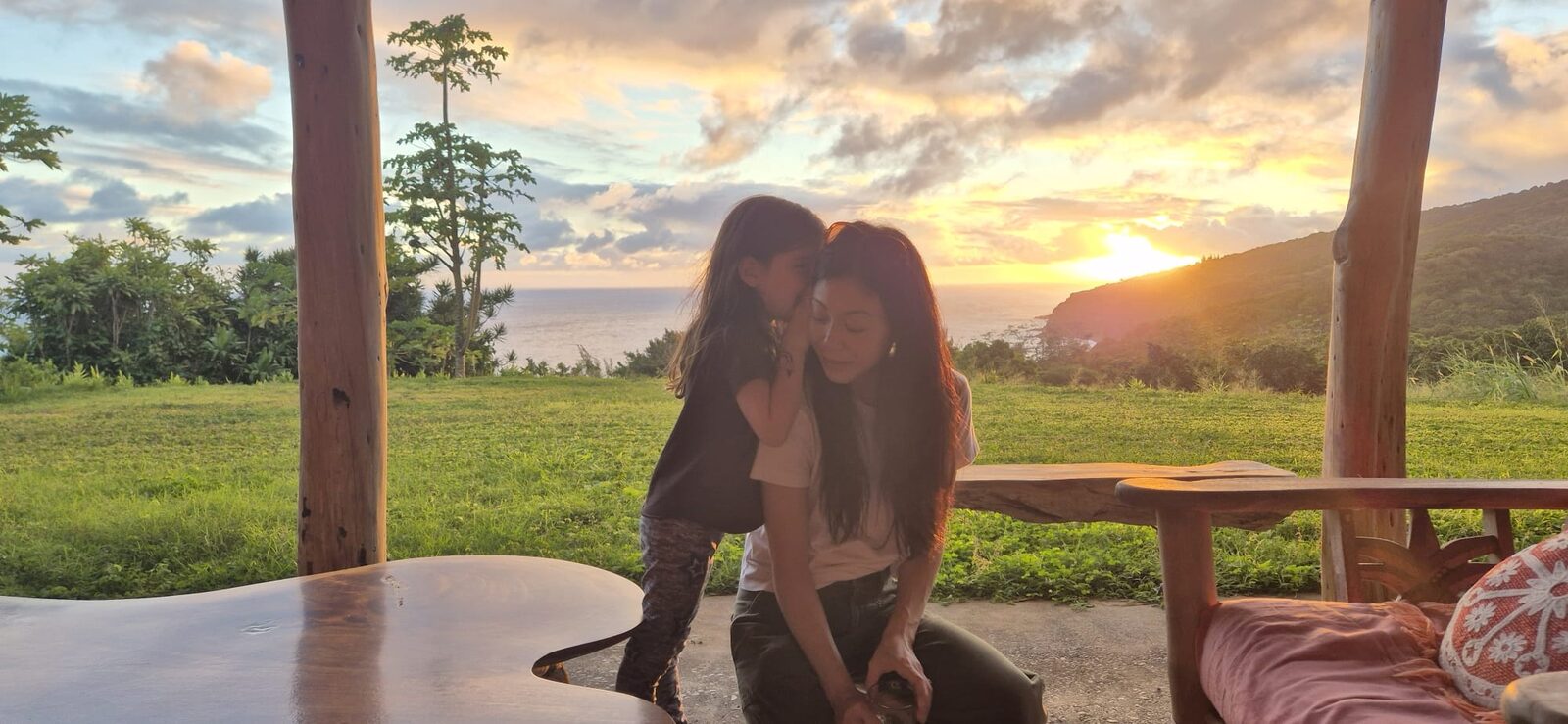 Brittany Ishibashi kneels on a covered patio at sunset, eyes closed and smiling, as her young daughter leans in to whisper in her ear, with green lawn, the ocean, and hills behind them.