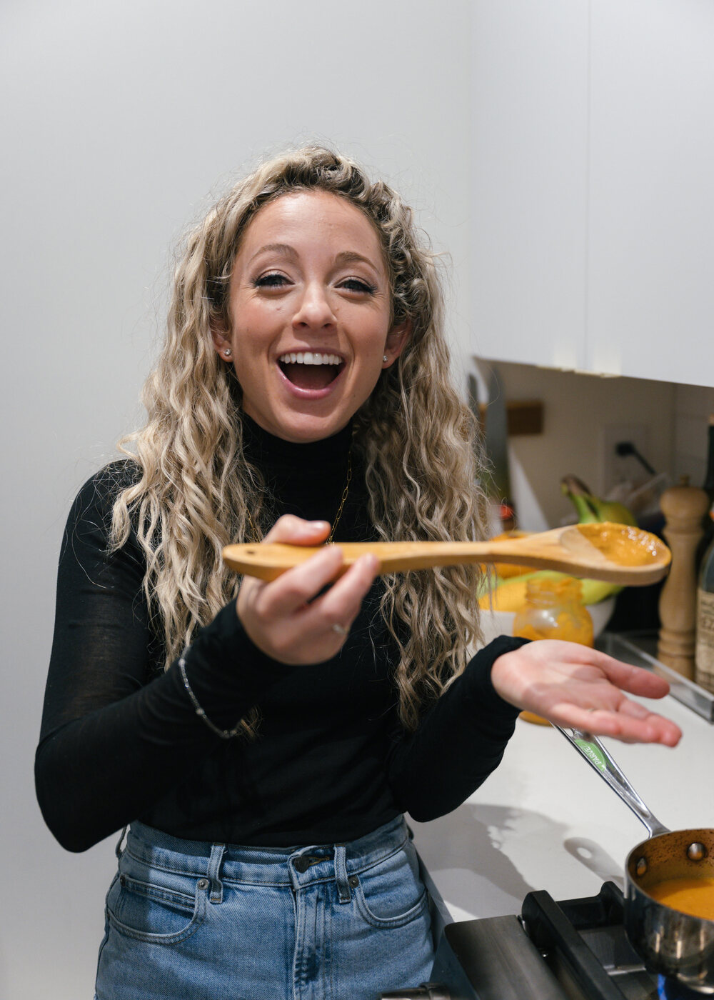 Cali Guttman laughs in her kitchen in a black turtleneck and jeans, holding out a wooden spoon of orange pur&eacute;e.