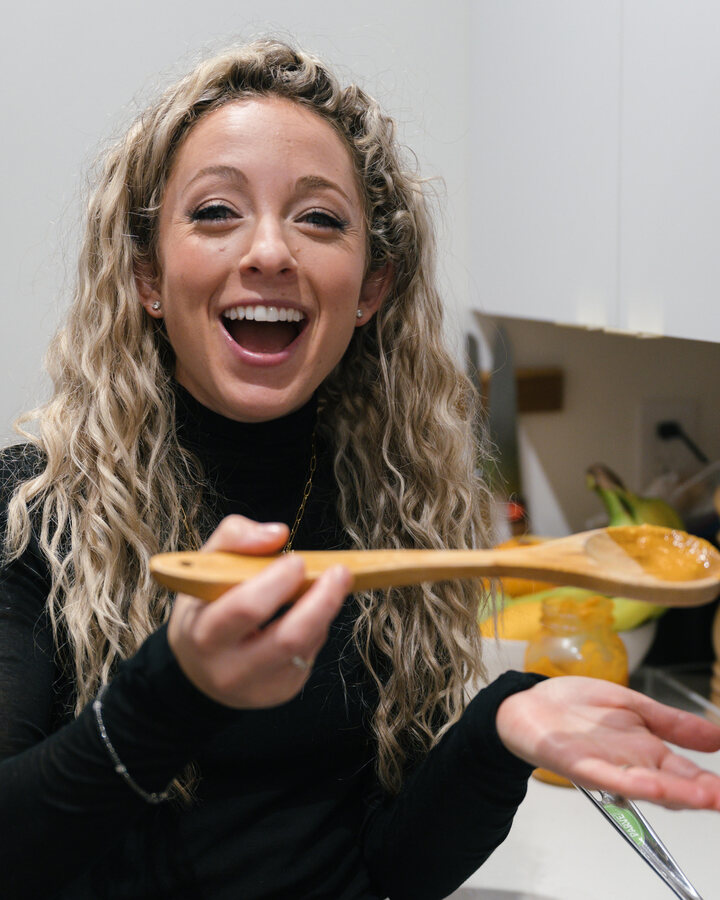 Cali Guttman laughs in her kitchen in a black turtleneck, holding out a wooden spoon of orange purée.
