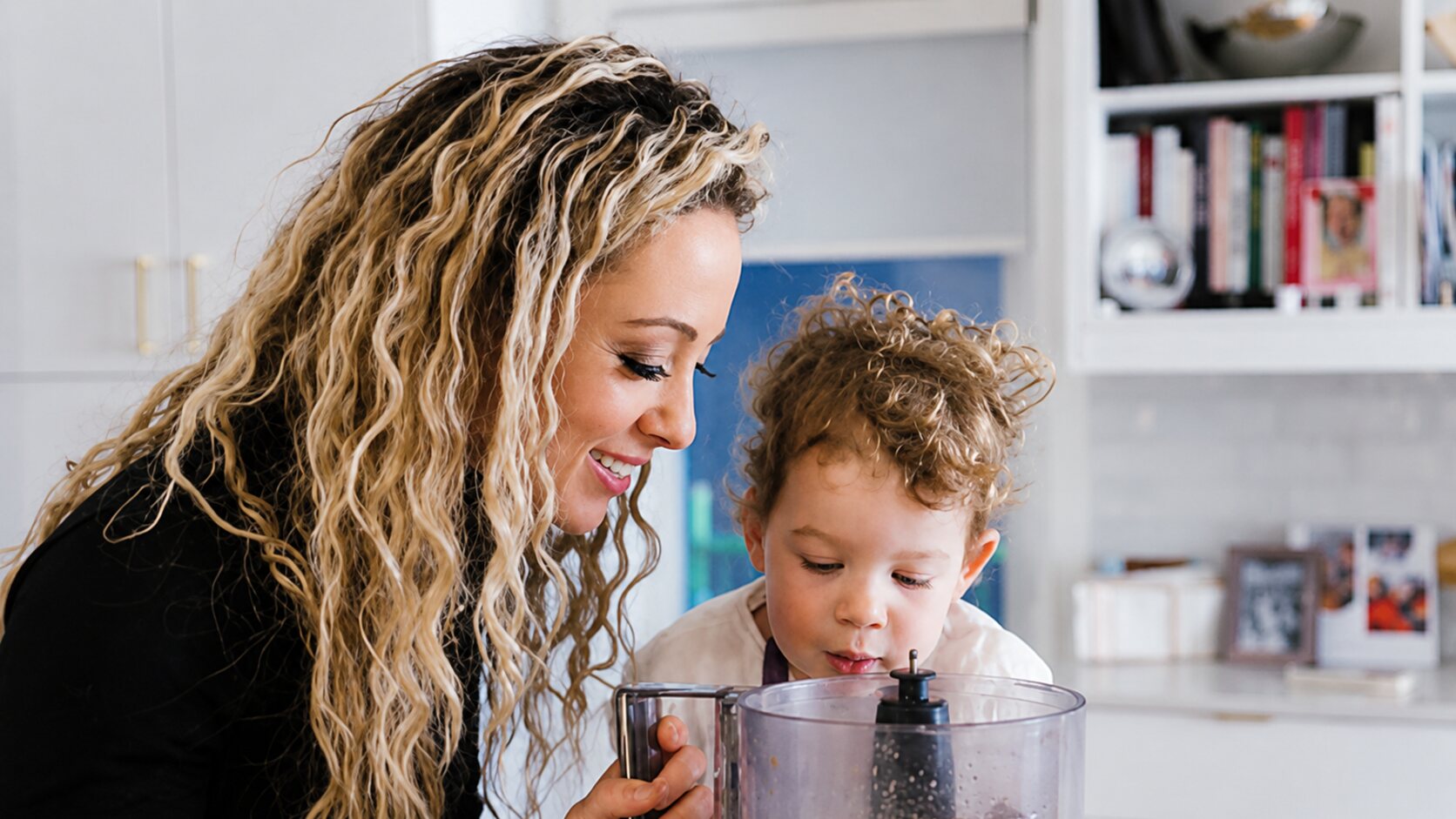 Cali Guttman leans over a kitchen counter beside her young son, both smiling down into a food processor bowl.