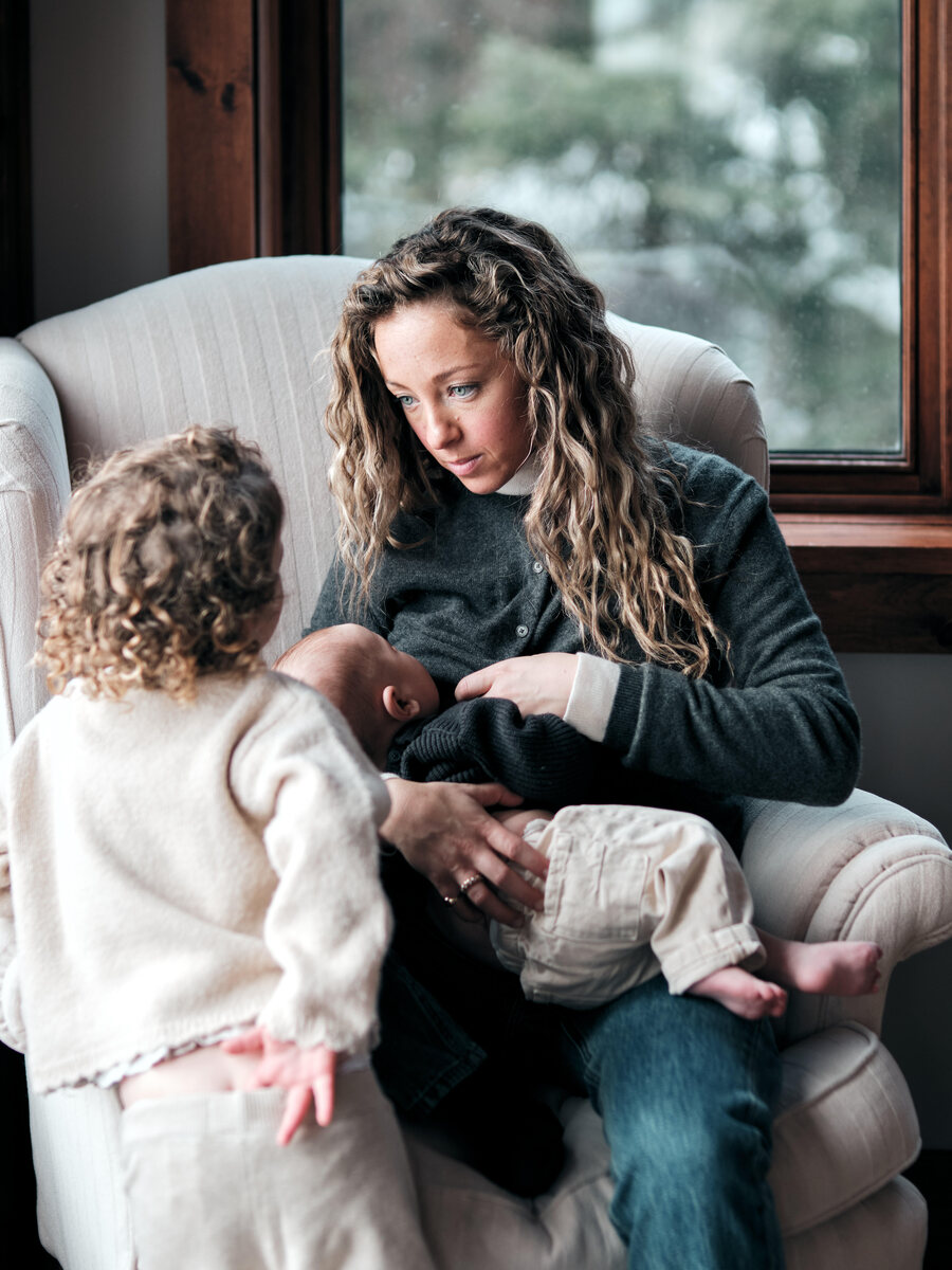 A mother sits in a pale armchair by a window nursing her baby while her toddler stands at her knee, watching.