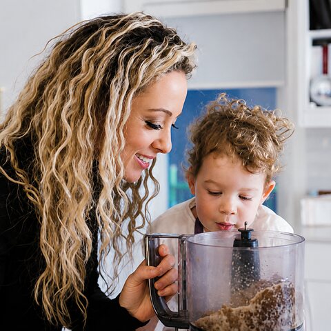 Cali Guttman leans over a kitchen counter beside her young son, both smiling down into a food processor bowl.