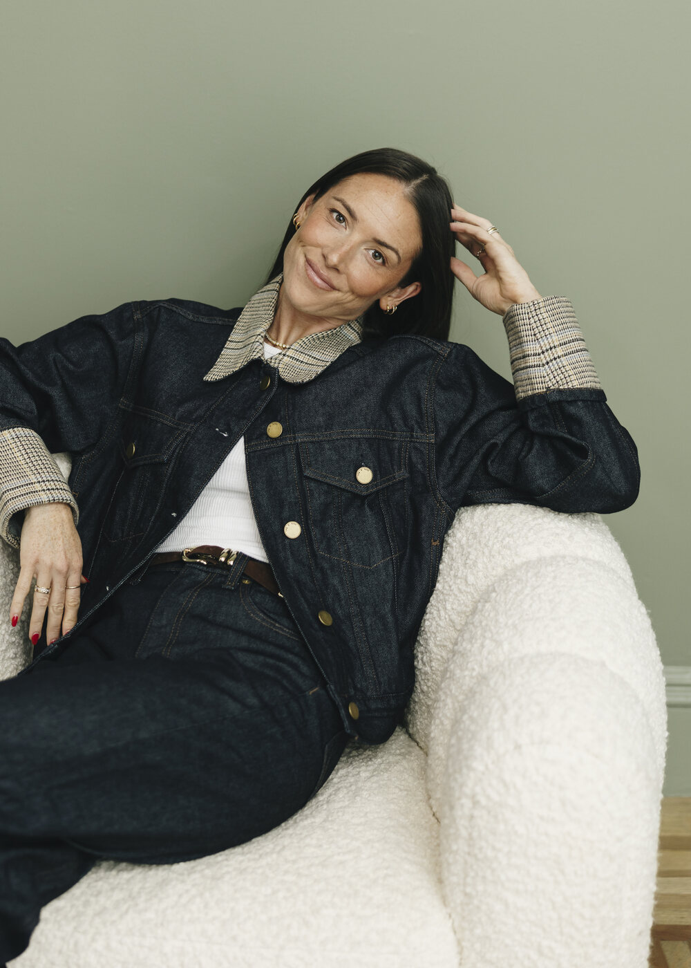 Cameron Rogers reclines in a cream bouclé chair in a denim jacket, one hand in her hair, smiling warmly at the camera.