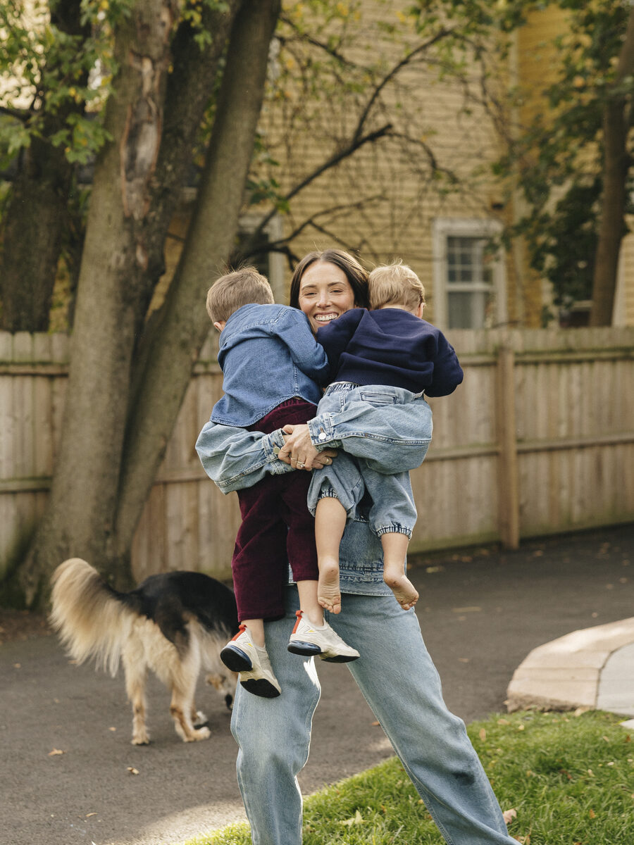 Cameron Rogers laughs while lifting both of her young sons in a joyful hug on a sunlit lawn, the family dog trotting past behind them.