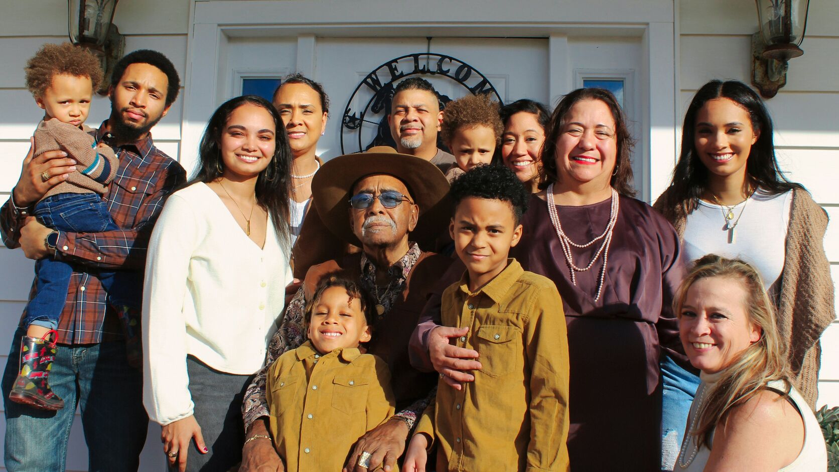 Three generations of a family gather on a white front porch beneath a wrought-iron Welcome sign, the grandfather in a brown cowboy hat seated at the centre with his grandsons in matching mustard corduroy shirts, everyone smiling in low autumn sun.