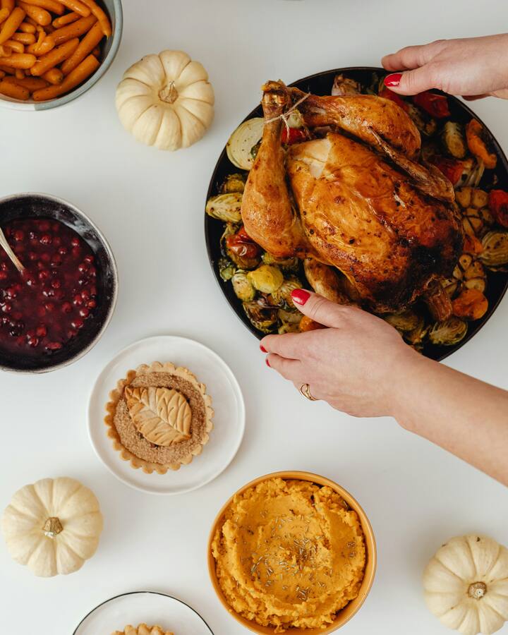 Overhead view of a Thanksgiving spread on a white table: hands with red nails set down a roast bird on a platter of Brussels sprouts and tomatoes, beside bowls of glazed carrots, cranberry sauce and mashed sweet potato, leaf-topped tarts and small white pumpkins.