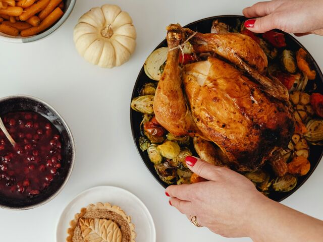 Overhead view of a Thanksgiving spread on a white table: hands with red nails set down a roast bird on a platter of Brussels sprouts and tomatoes, beside bowls of glazed carrots, cranberry sauce and mashed sweet potato, leaf-topped tarts and small white pumpkins.