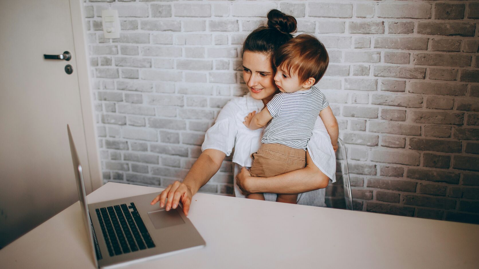 A smiling mother in a white blouse holds her toddler on her hip while reaching for a laptop at a white desk against an exposed brick wall.