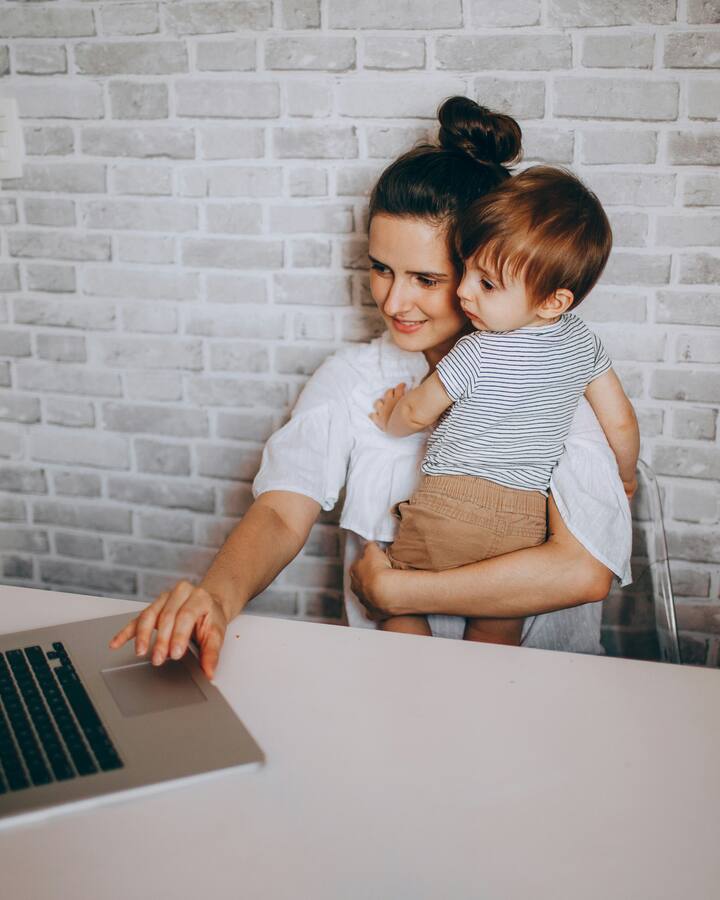 A smiling mother in a white blouse holds her toddler on her hip while reaching for a laptop at a white desk against an exposed brick wall.