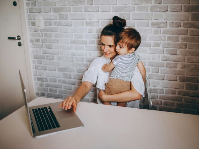 A smiling mother in a white blouse holds her toddler on her hip while reaching for a laptop at a white desk against an exposed brick wall.