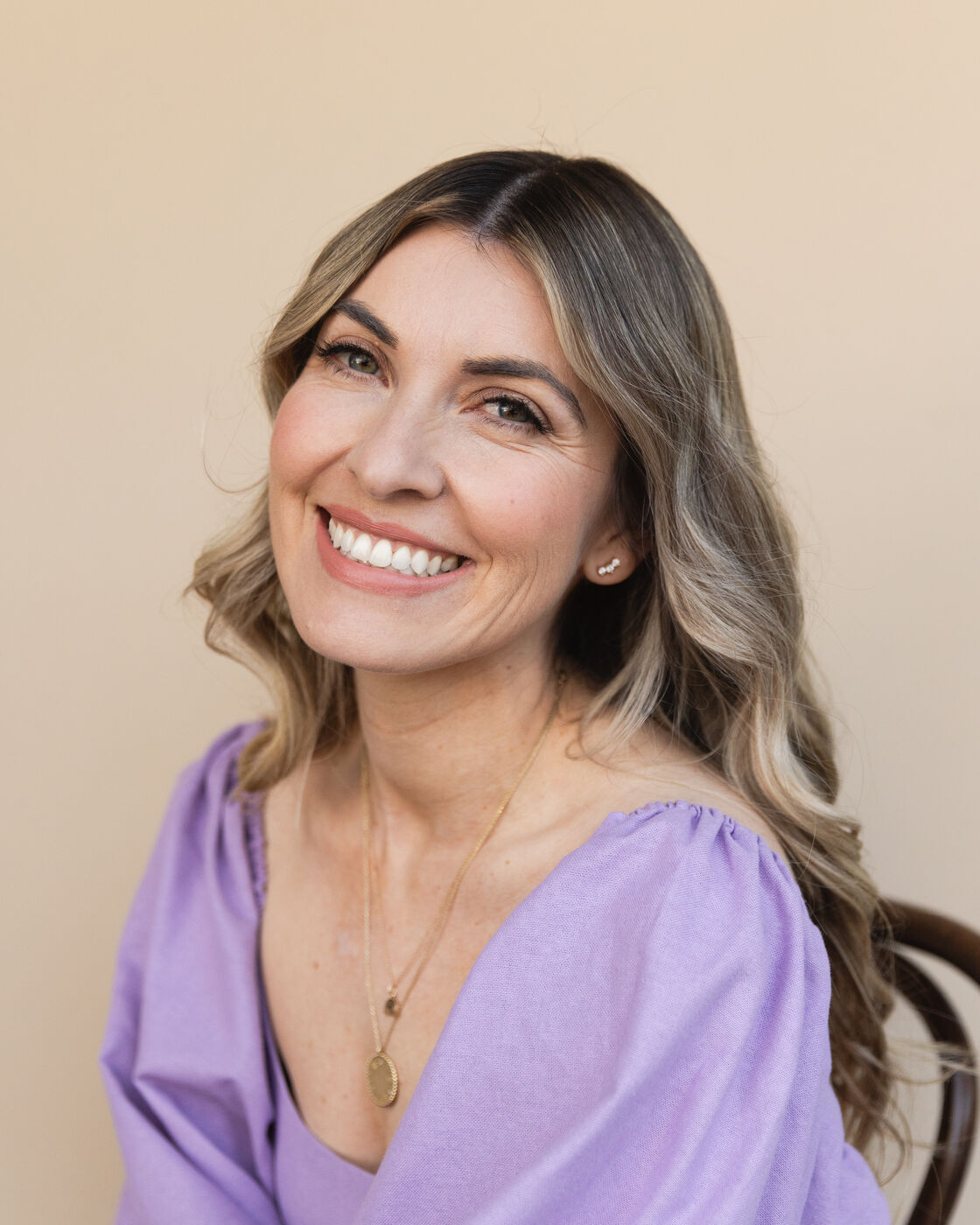 Dr. Cassidy Freitas, in a lilac puff-sleeve top and a gold pendant necklace, smiles at the camera with her head tilted against a warm beige backdrop.
