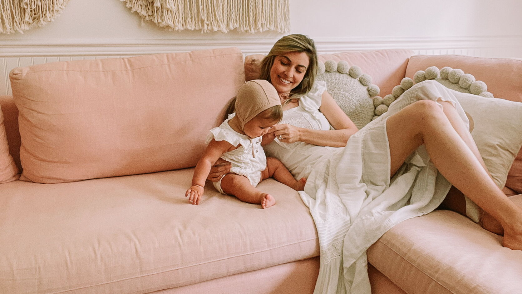 Dr. Cassidy Freitas, in a white dress, smiles down at a baby in a white ruffled romper and knit bonnet as they sit together on a blush-pink sofa beneath a cream macramé wall hanging.
