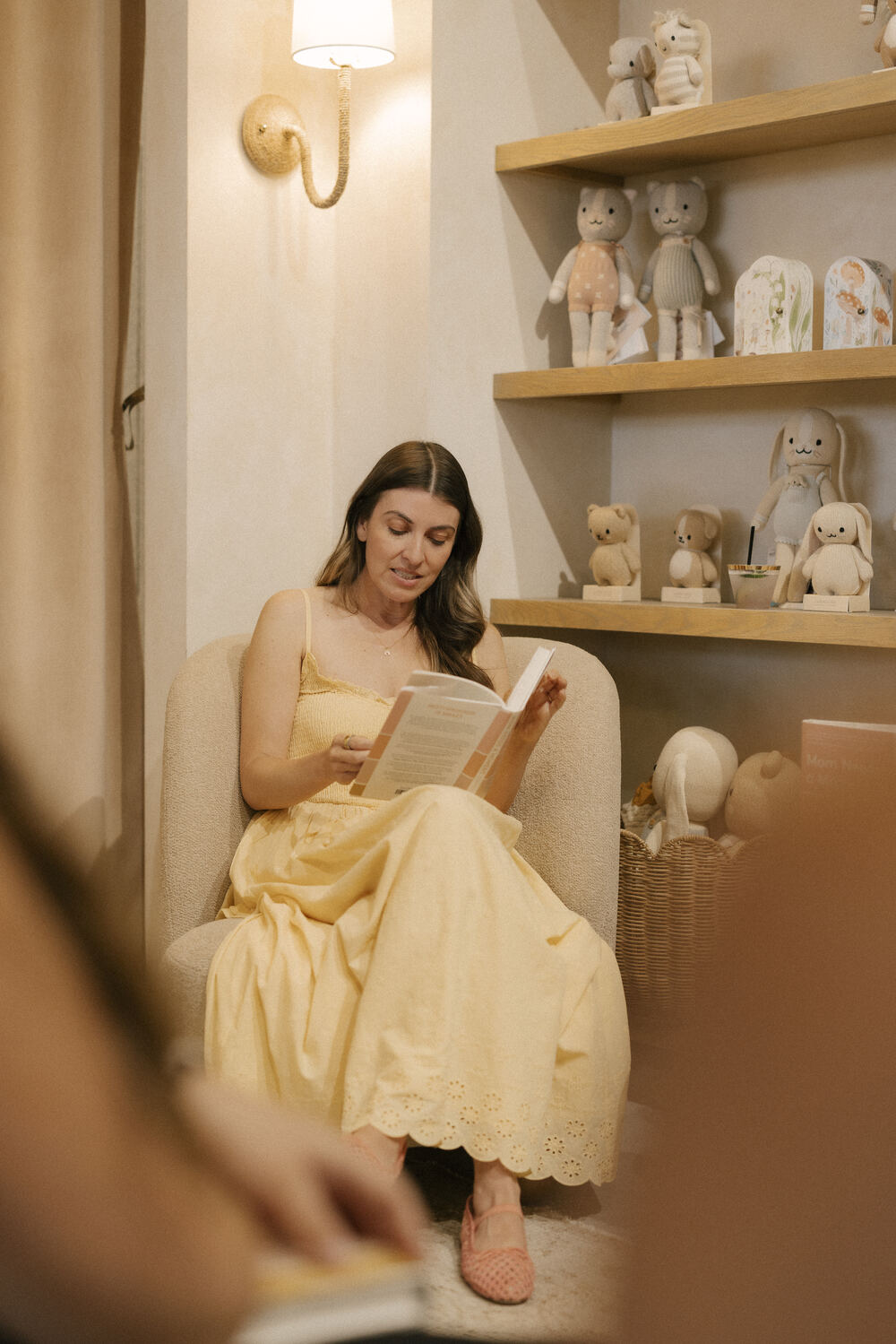 Dr. Cassidy Freitas, in a pale yellow eyelet sundress, reads a book in a cream armchair in a softly lit nursery corner, wooden shelves of knitted stuffed animals behind her.