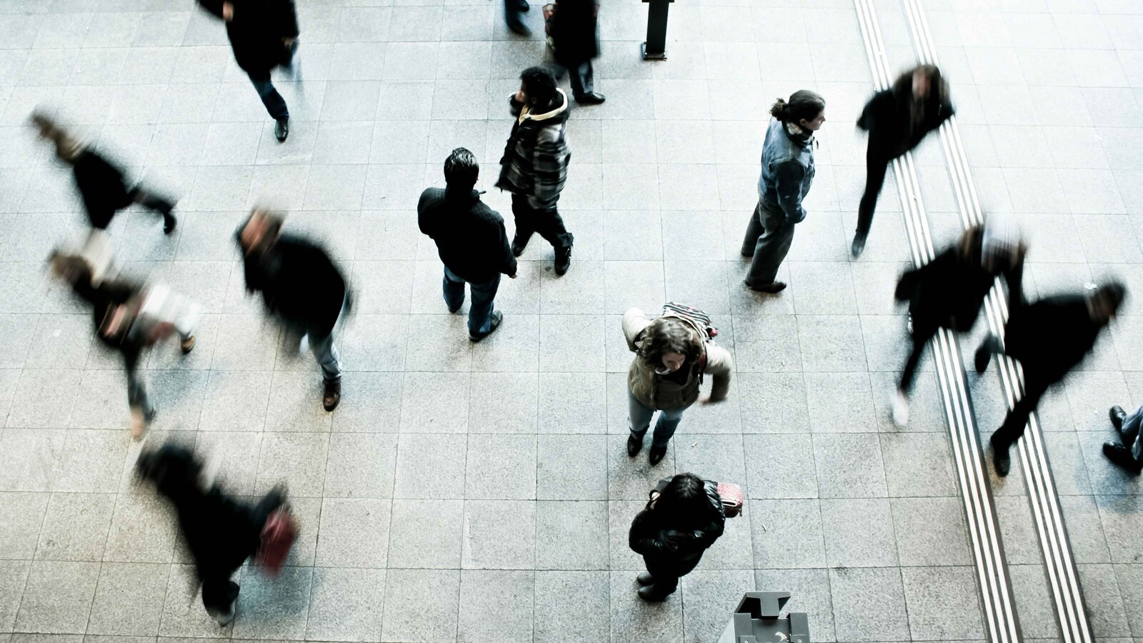 A high overhead view of a crowd of people walking across a tiled public plaza, their movement blurred.