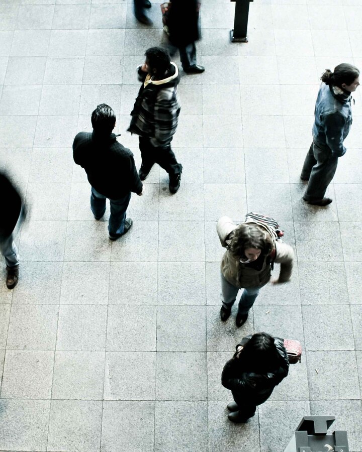 A high overhead view of a crowd of people walking across a tiled public plaza, their movement blurred.