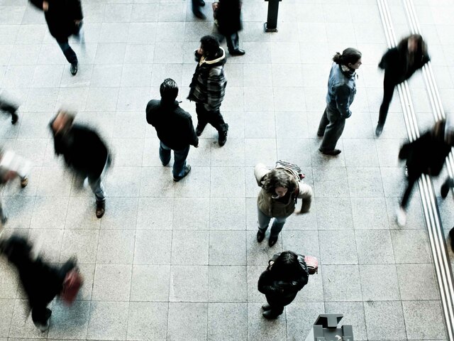 A high overhead view of a crowd of people walking across a tiled public plaza, their movement blurred.