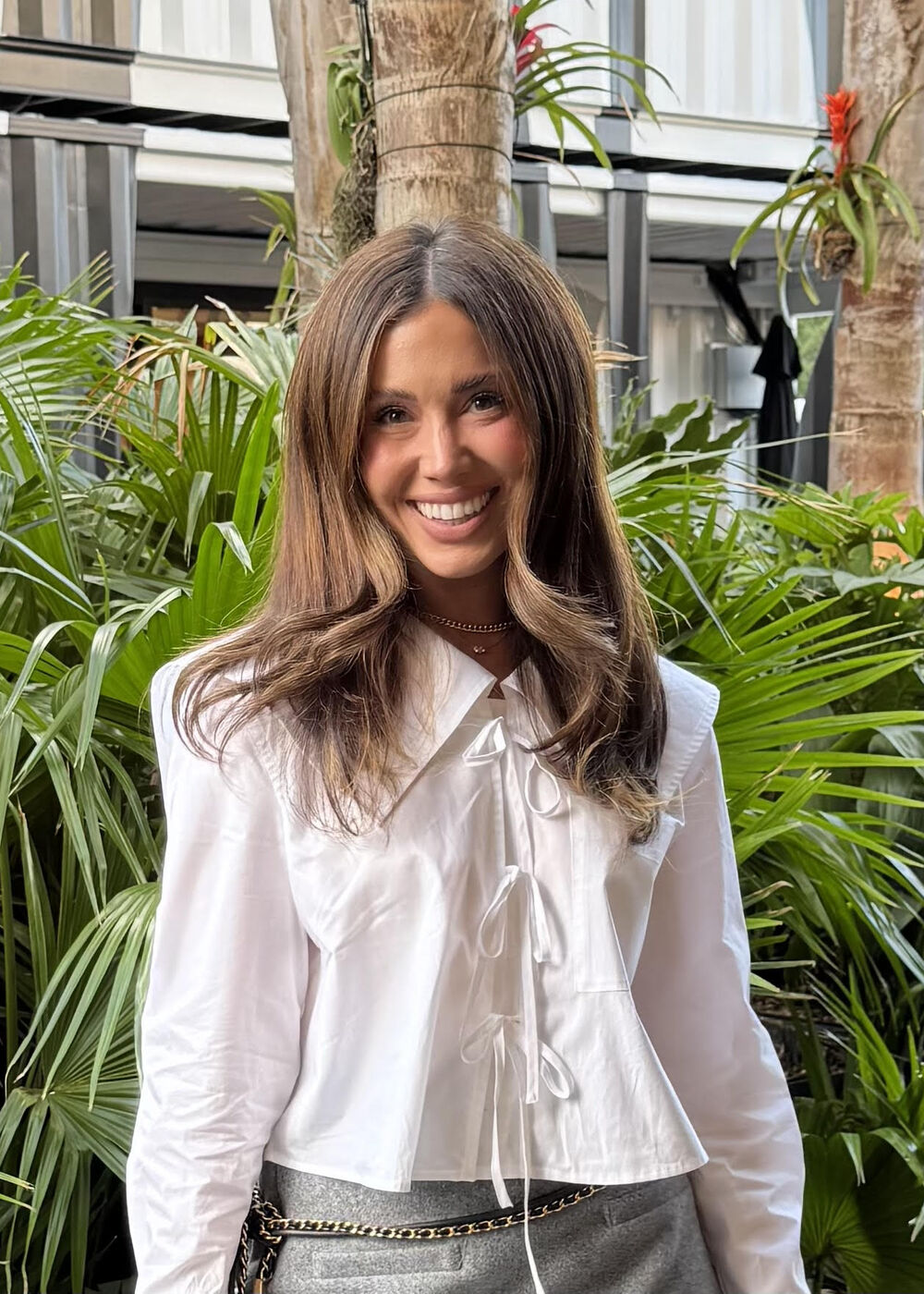 Cecily Bauchmann smiles in a white bow-trimmed blouse and grey trousers, standing in front of a wall of palm fronds.