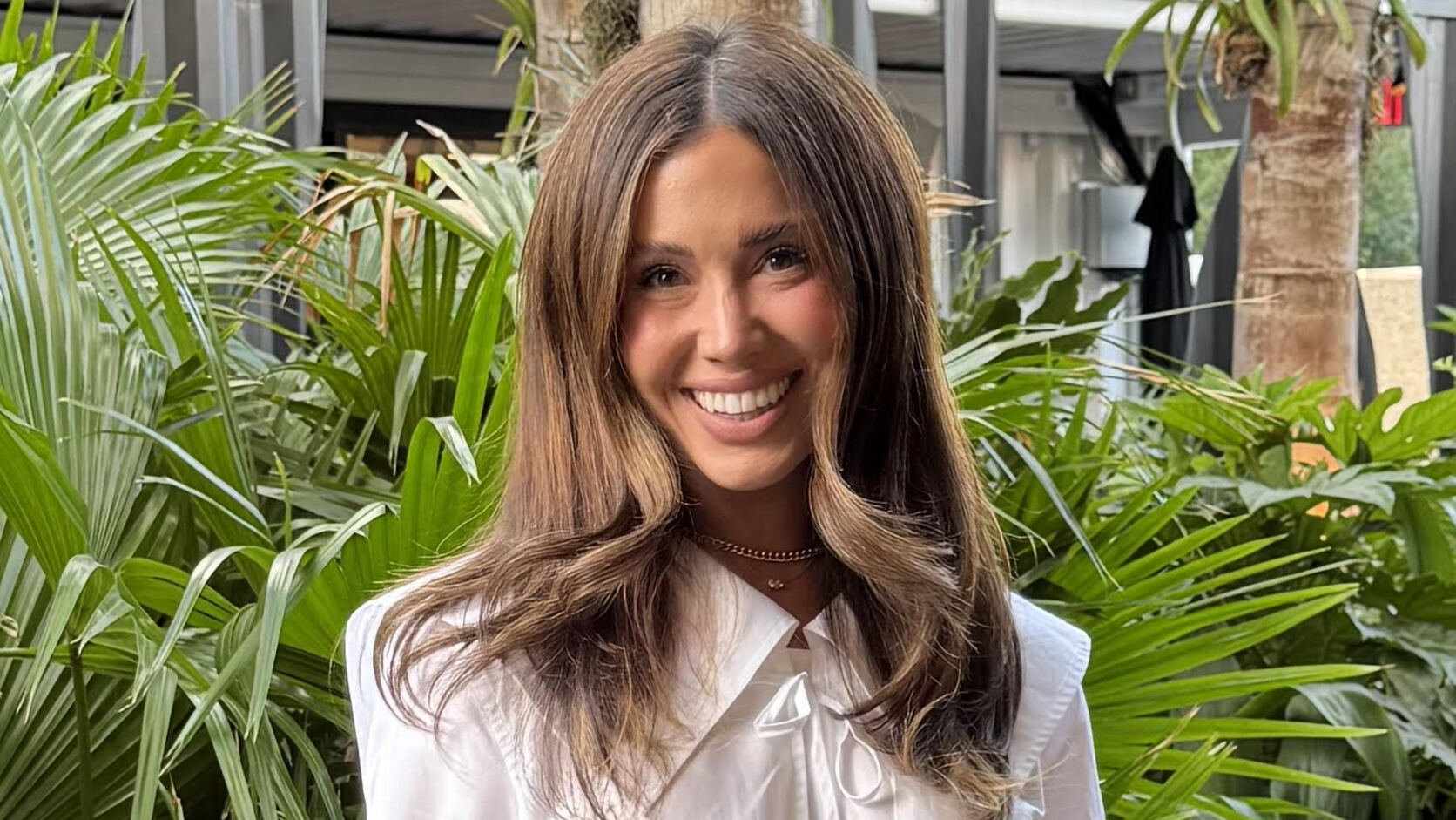 Cecily Bauchmann smiles in a white bow-trimmed blouse and grey trousers, standing in front of a wall of palm fronds.