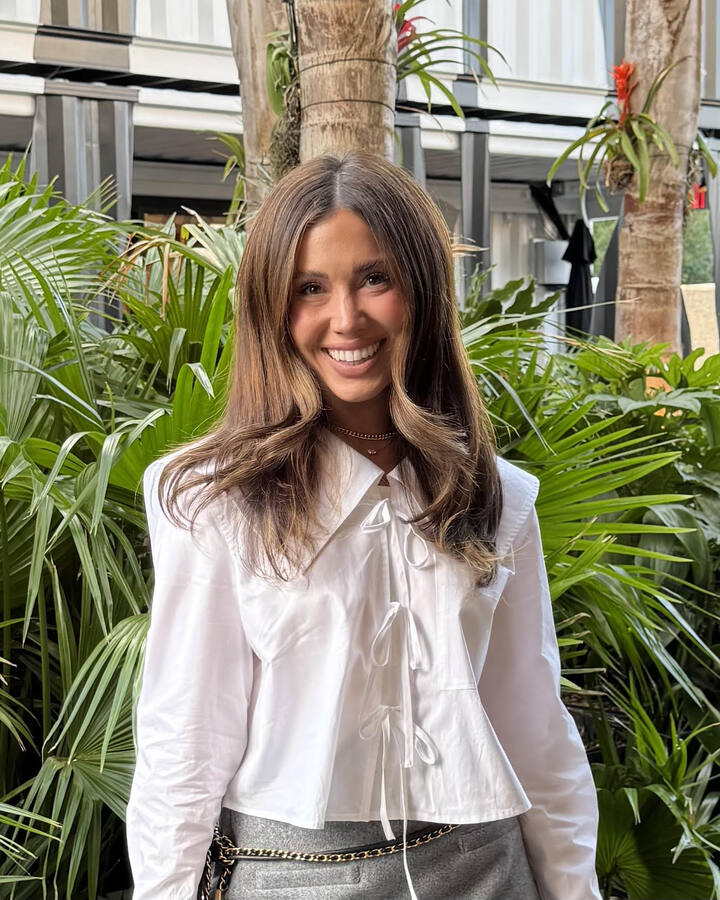 Cecily Bauchmann smiles in a white bow-trimmed blouse and grey trousers, standing in front of a wall of palm fronds.